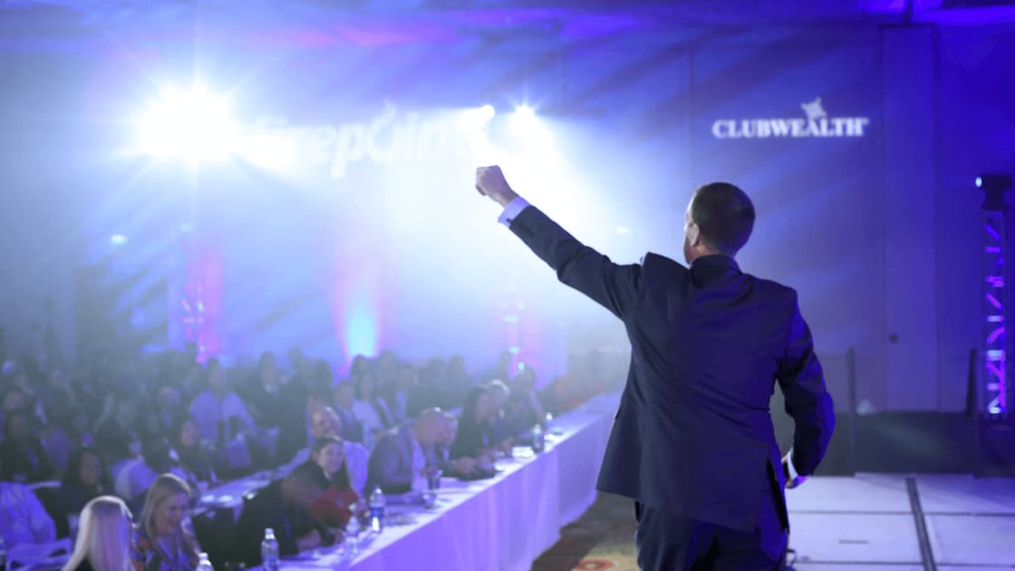 A speaker on stage in front of a full room at a Club Wealth live event